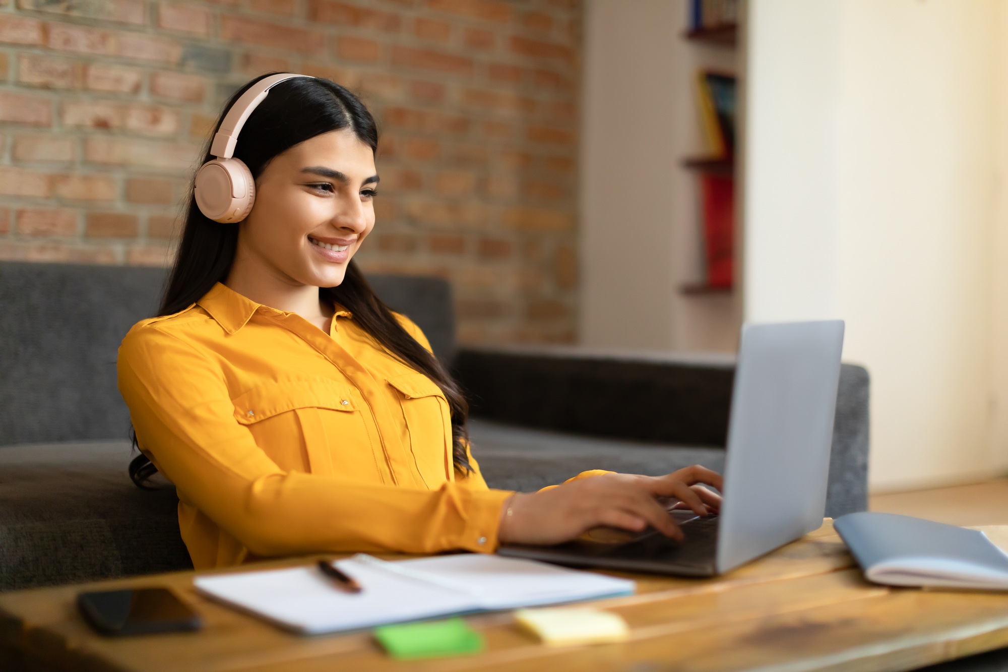Smiling young spanish lady wearing wireless headphones and using laptop at home, studying or working